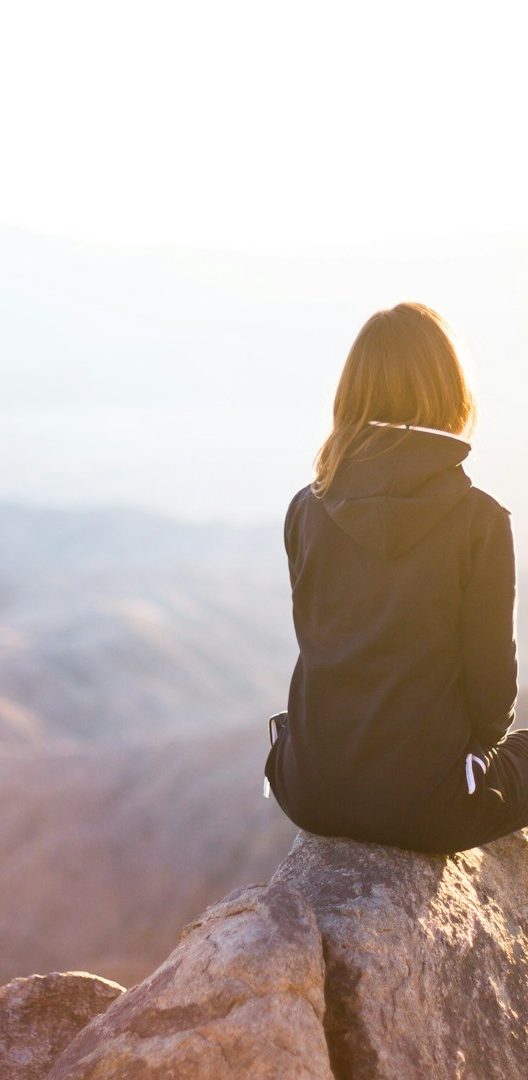 Medical Coaching Eine Person sitzt auf einem Felsen und blickt in die Ferne bei Sonnenlicht.