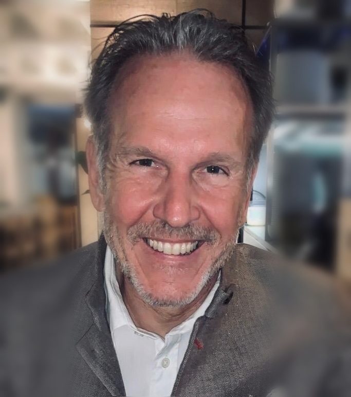 Mann mit grauen Haaren und lächelndem Gesicht, trägt einen grauen Pullover.